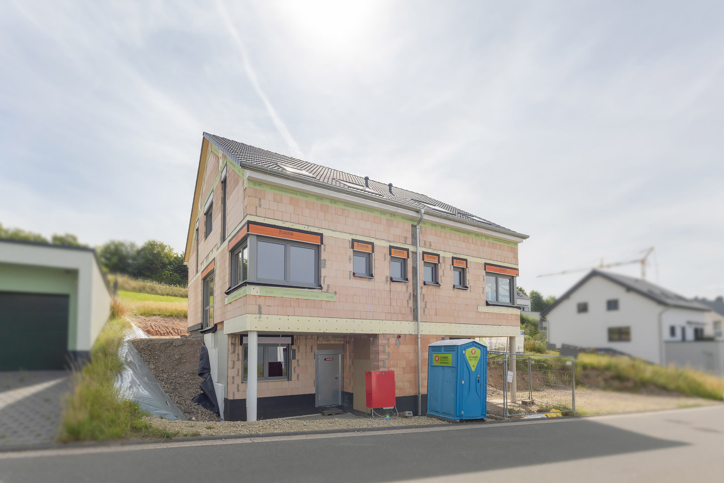 Ein im Bau befindliches Mehrfamilienhaus im Rohbauzustand auf einer Baustelle. Das Foto zeigt die unfertige Fassade aus rotem Ziegelmauerwerk, bereits eingesetzte dunkelgraue Fenster und ein fertig gedecktes Dach.